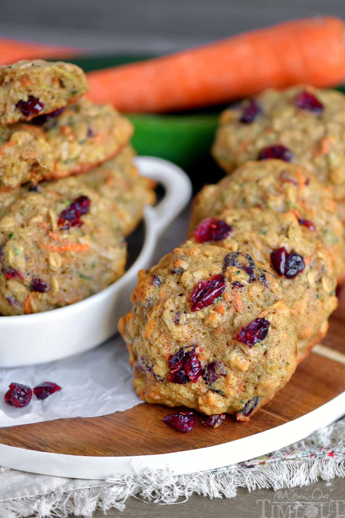 Zucchini oatmeal cookies in a row on a wood board.  Craisins can be seen on top of the cookies along with a carrot and zucchini in the background.