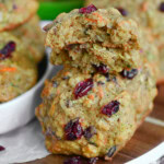 Zucchini cookies on a wood board with white rim. Top cookie has been halved. Carrot and zucchini can be seen in the background.