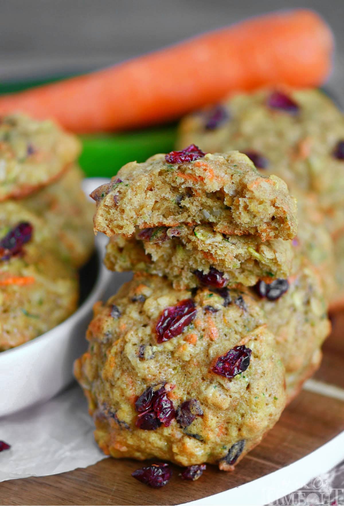 Zucchini cookies on a wood board with white rim. Top cookie has been halved. Carrot and zucchini can be seen in the background.