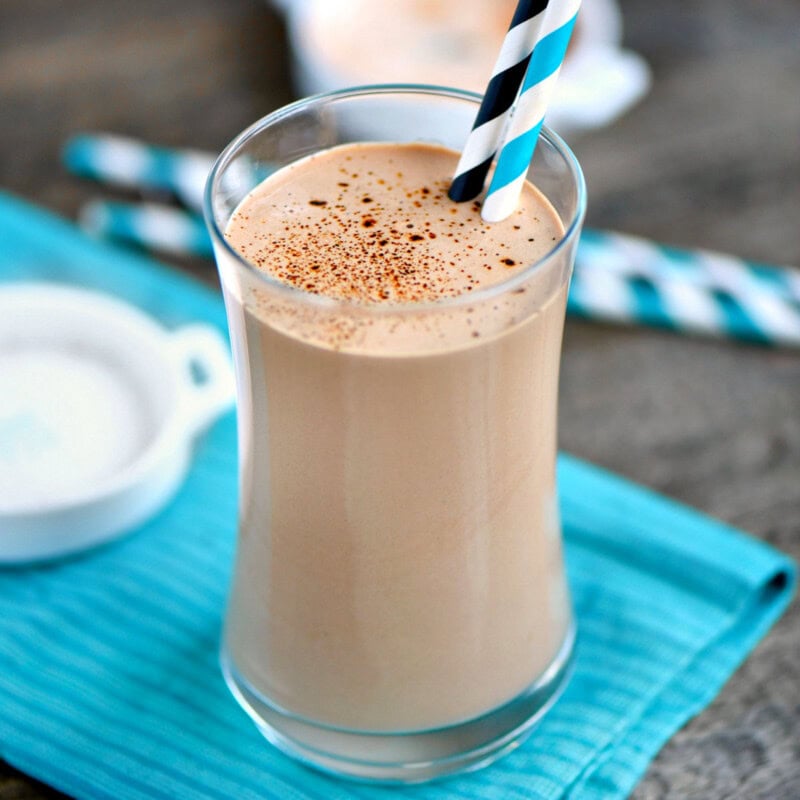 Coffee protein shake in a tall glass with two paper straws. Glass is sitting on teal napkin on a wood surface.