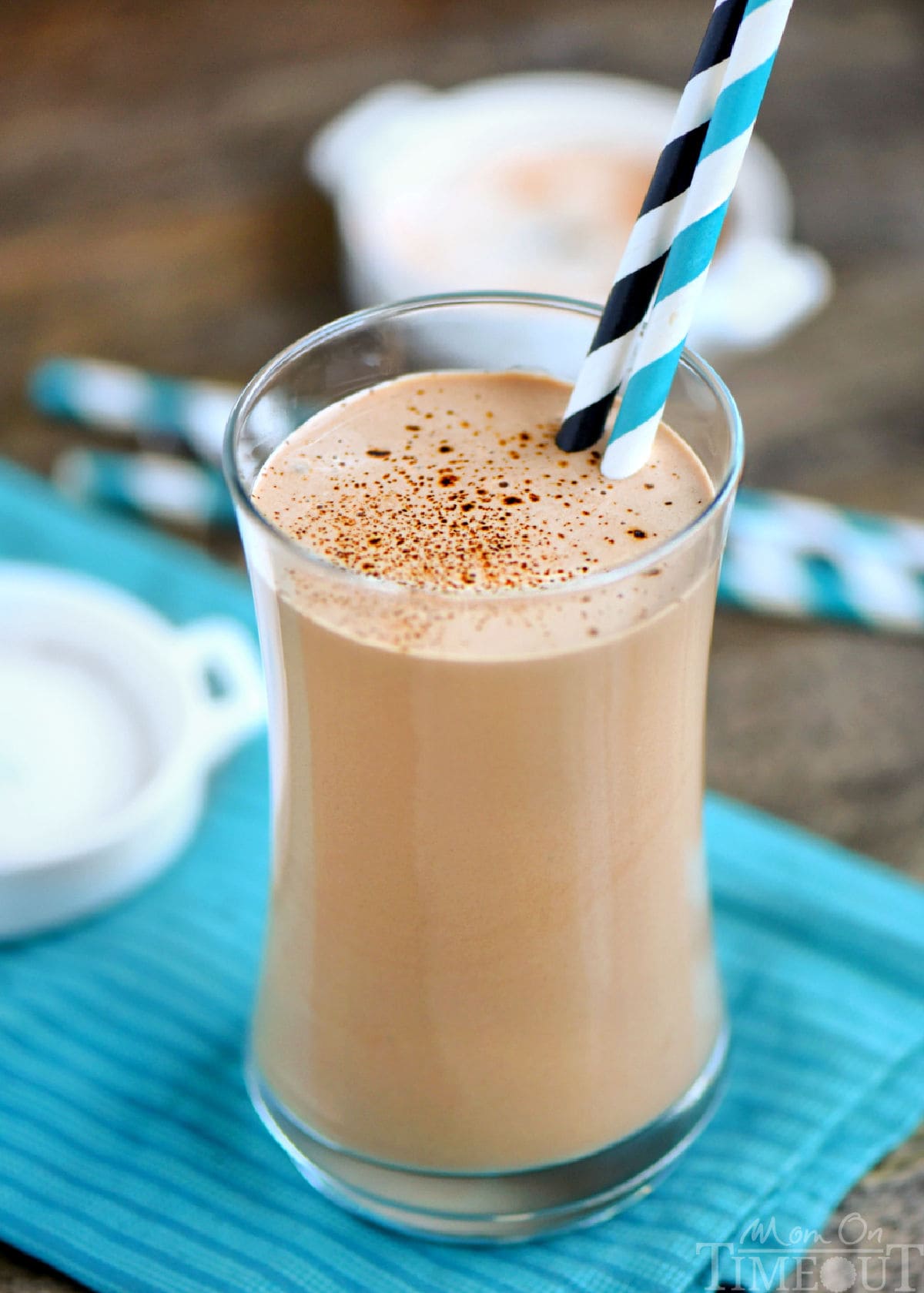 Coffee protein shake in a tall glass with two paper straws. Glass is sitting on teal napkin on a wood surface.