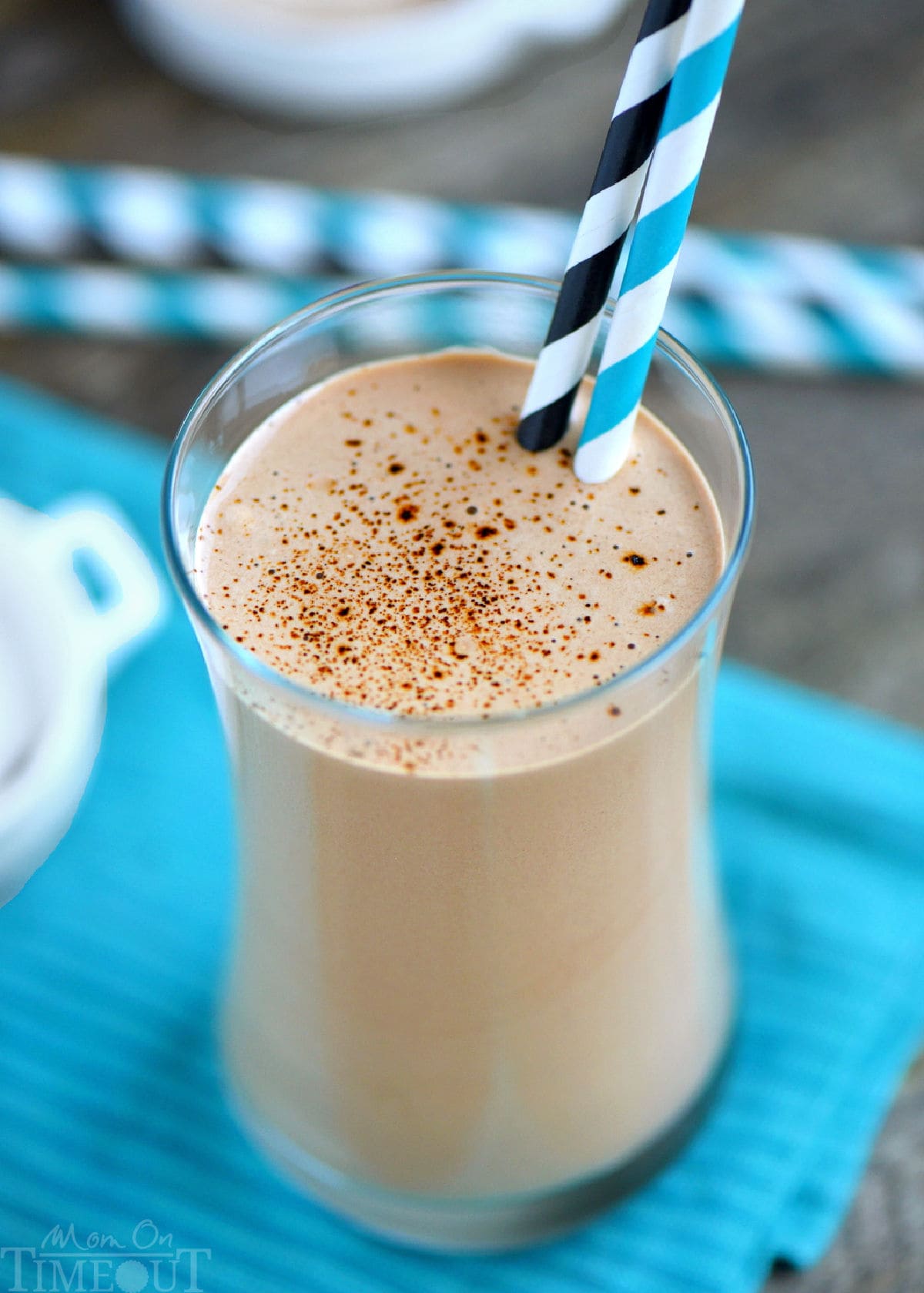 Angled top down view of protein and coffee shake poured into a tall glass and finished off with a dusting of espresso powder. Shake is resting on a striped teal towel in front of a few straws.