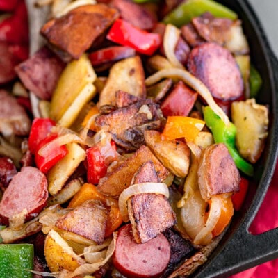 Kielbasa and potatoes in a cast iron skillet ready to be enjoyed. Skillet is resting on a red napkin. Peppers, onions, potatoes and kielbasa are all cut into bite sized pieces.