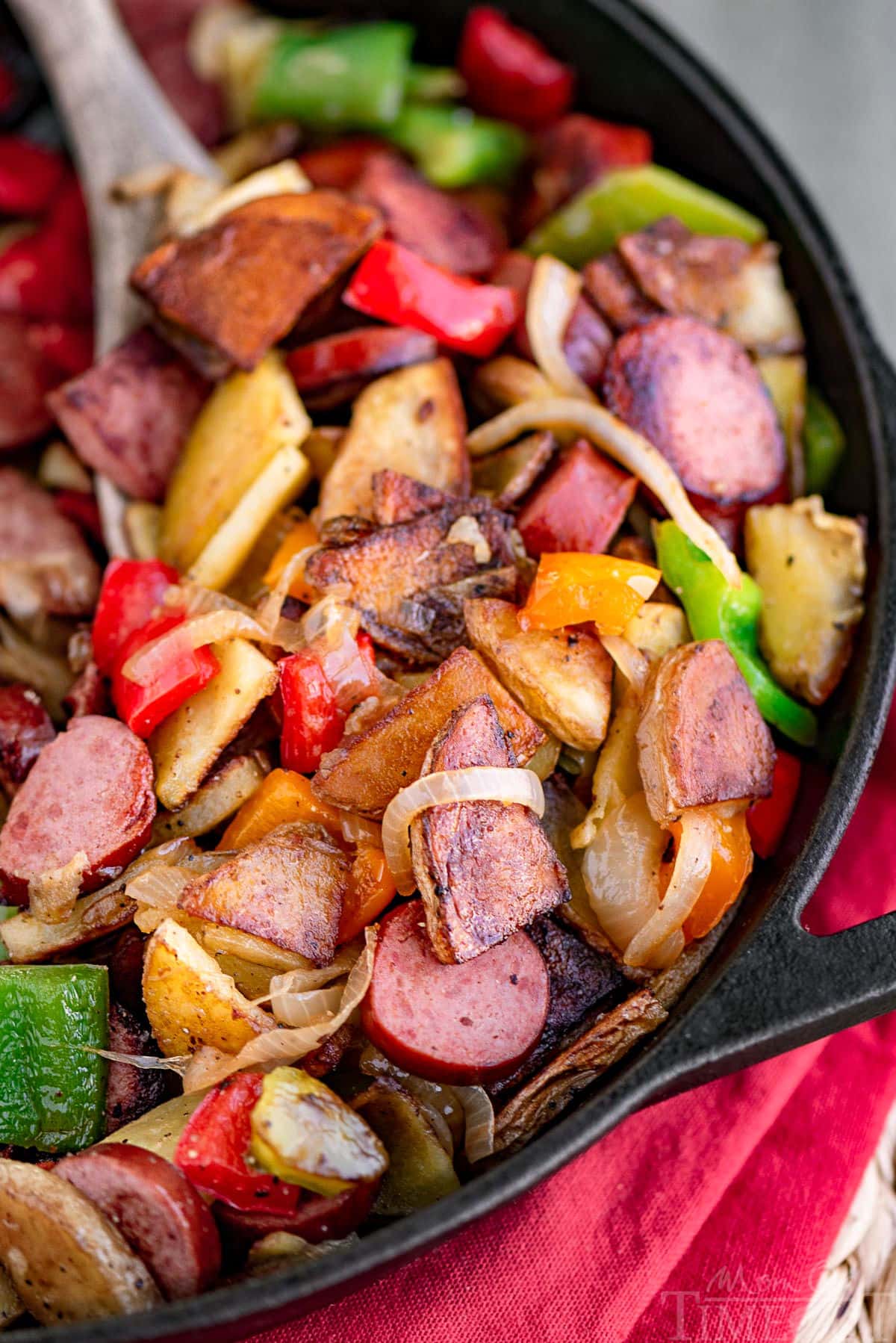 Kielbasa and potatoes in a cast iron skillet ready to be enjoyed. Skillet is resting on a red napkin. Peppers, onions, potatoes and kielbasa are all cut into bite sized pieces.