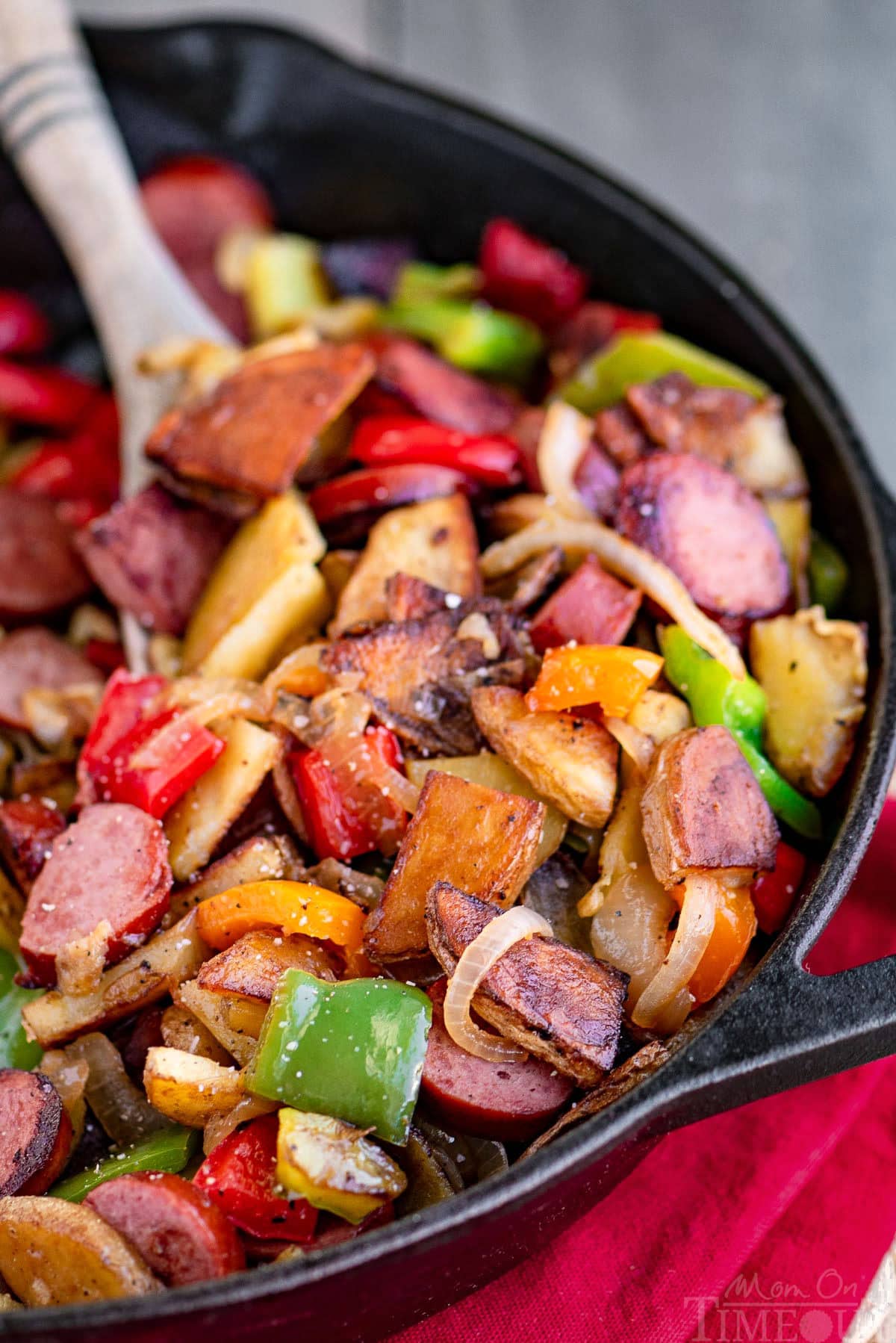 Kielbasa and potatoes in a cast iron skillet ready to be enjoyed. Skillet is resting on a red napkin. Peppers, onions, potatoes and kielbasa are all cut into bite sized pieces.