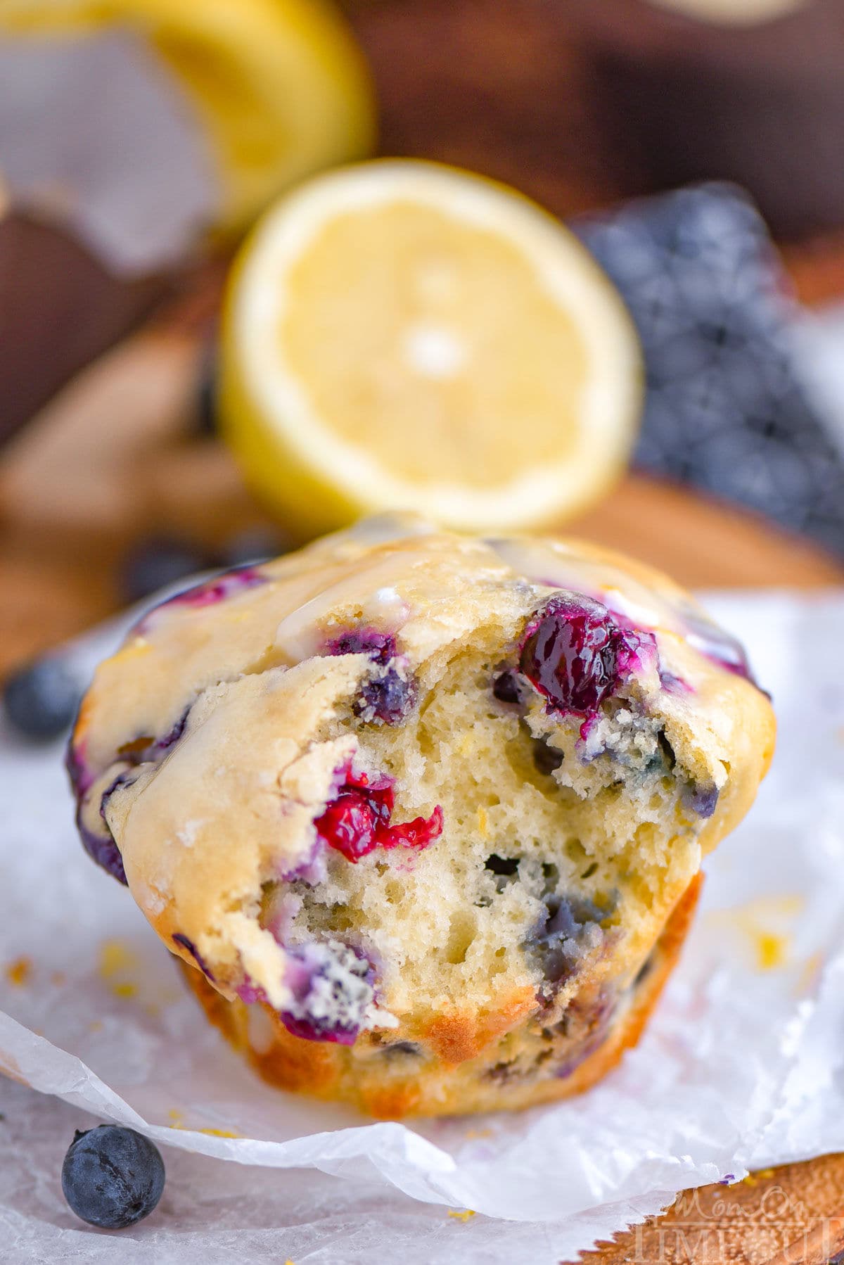 Blueberry muffin made with cream cheese and lemon sitting on white parchment with a bite taken out of the muffin. A lemon half can be seen in the background.