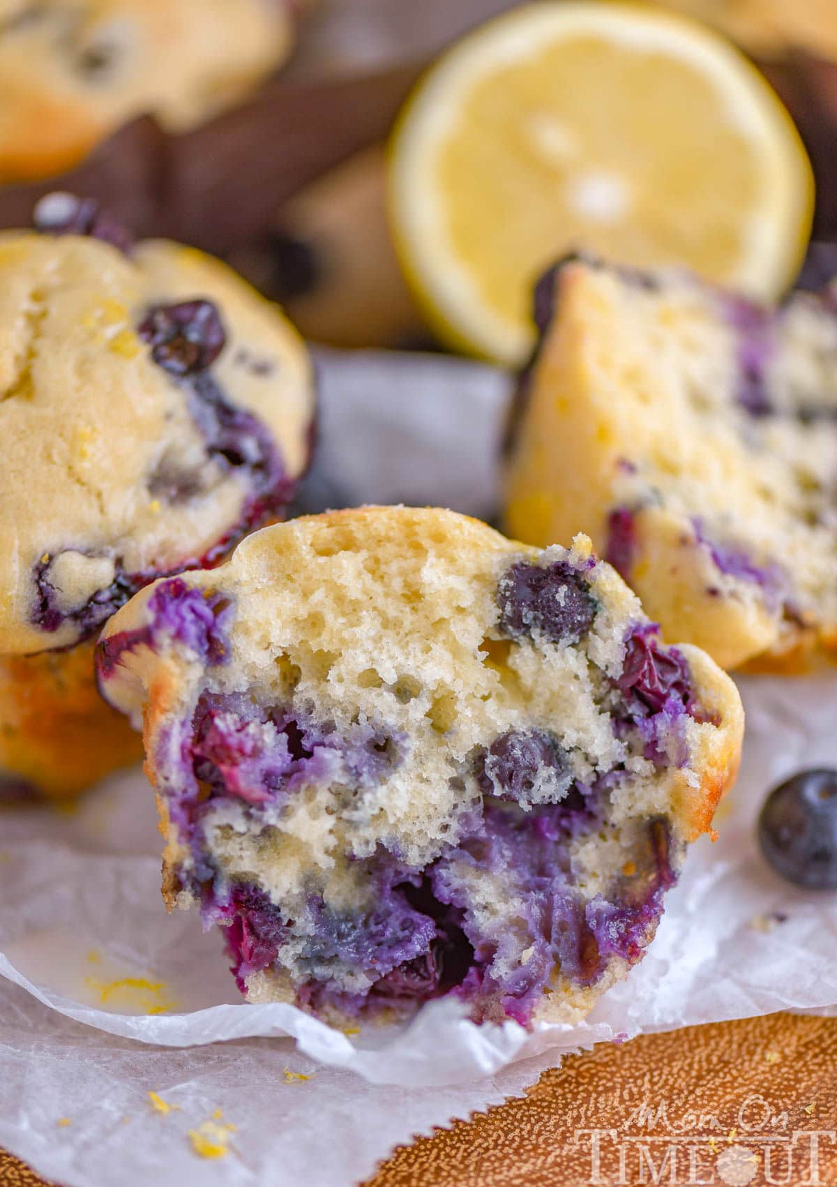 Blueberry Lemon Muffin split in half sitting on white parchment with more muffins and lemon halves in the background.