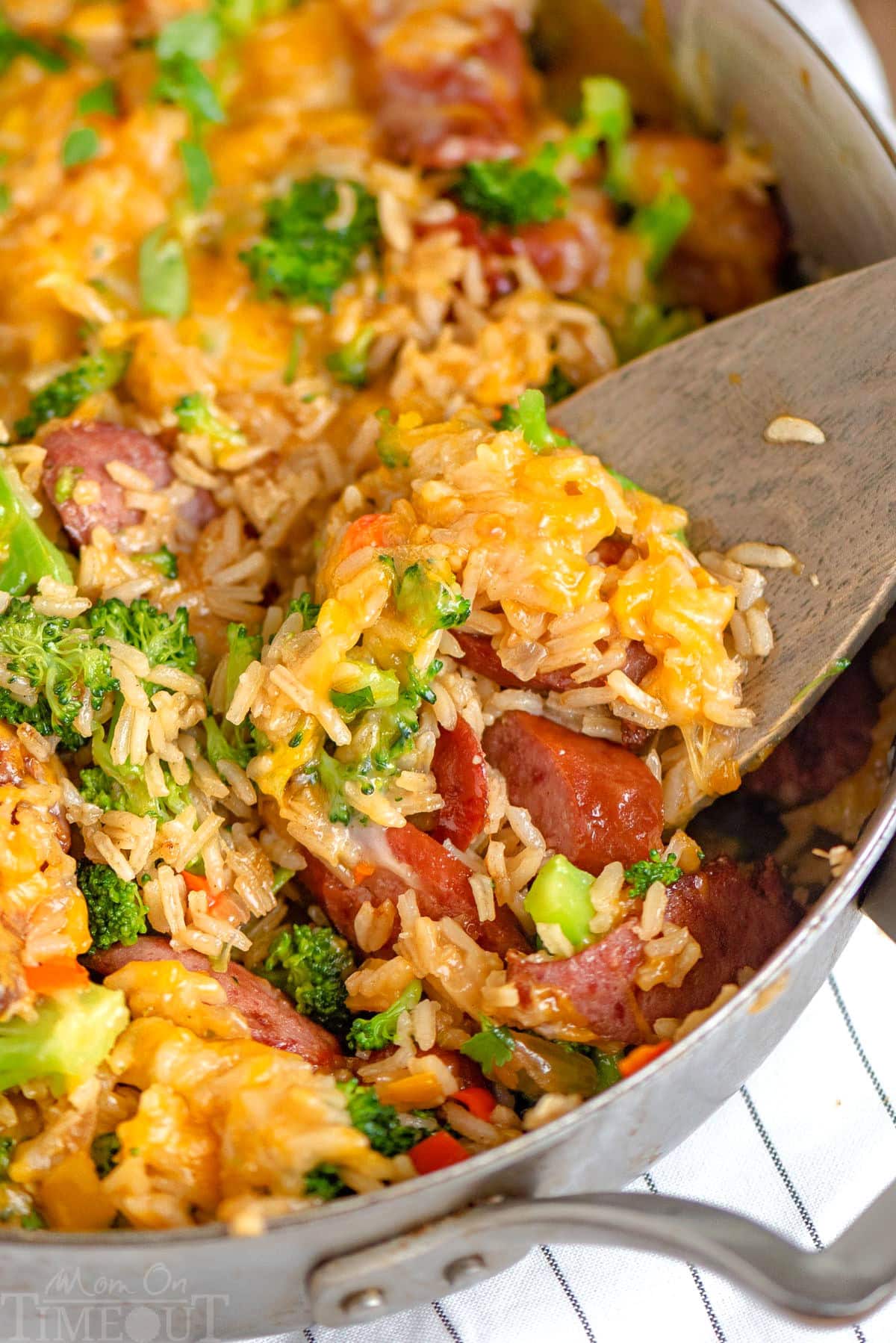 Wood spoon scooping out kielbasa skillet with rice and broccoli.