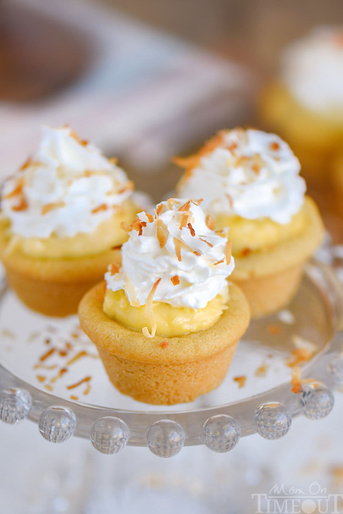 Three coconut cream pie cookies cups on glass stand with whipped cream and toasted coconut on top.