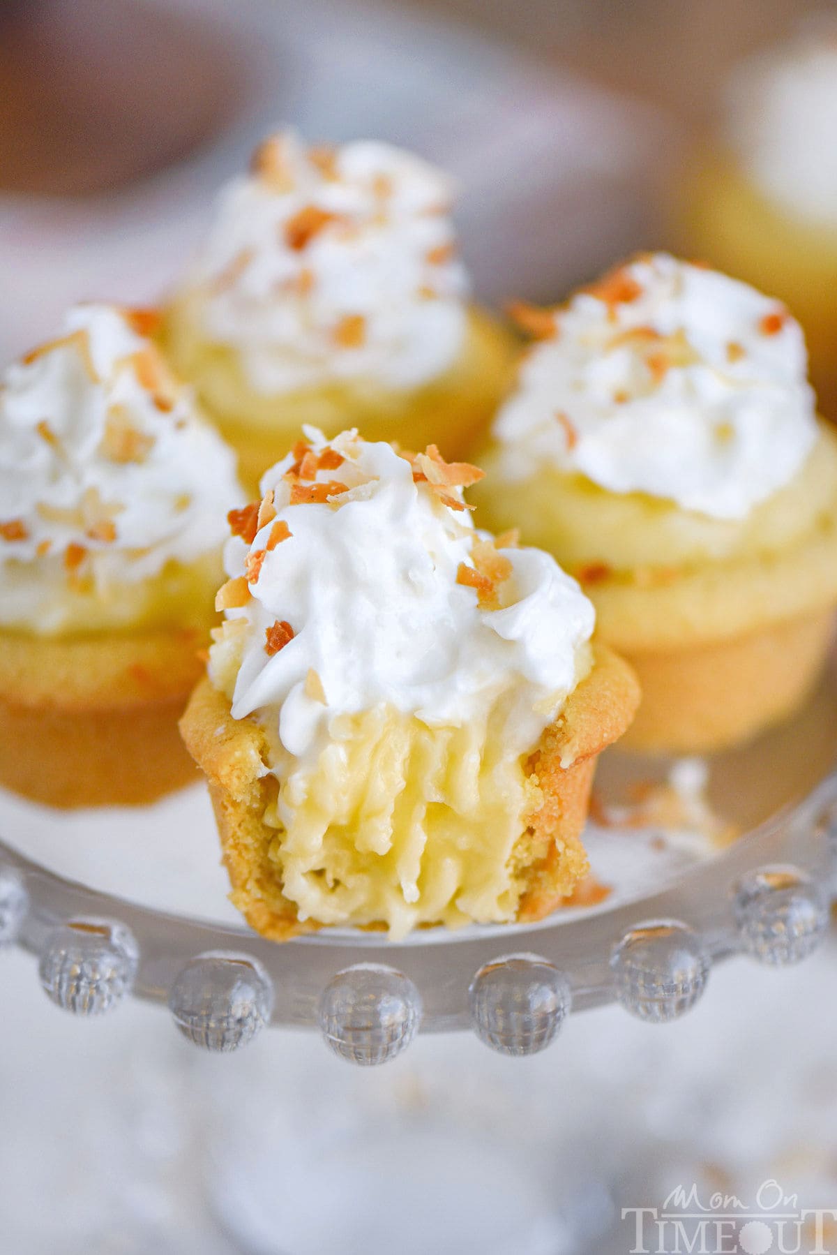 Four mini coconut cream pies on glass cake stand topped with whipped cream and toasted coconut. The front one has a bite taken out of it.