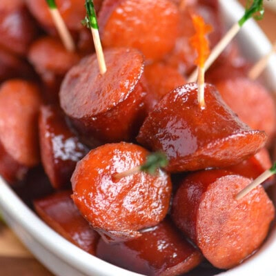 Pieces of kielbasa have been cooked in a slow cooker in a mouthwatering sauce and are shown in a white bowl with toothpicks in them.