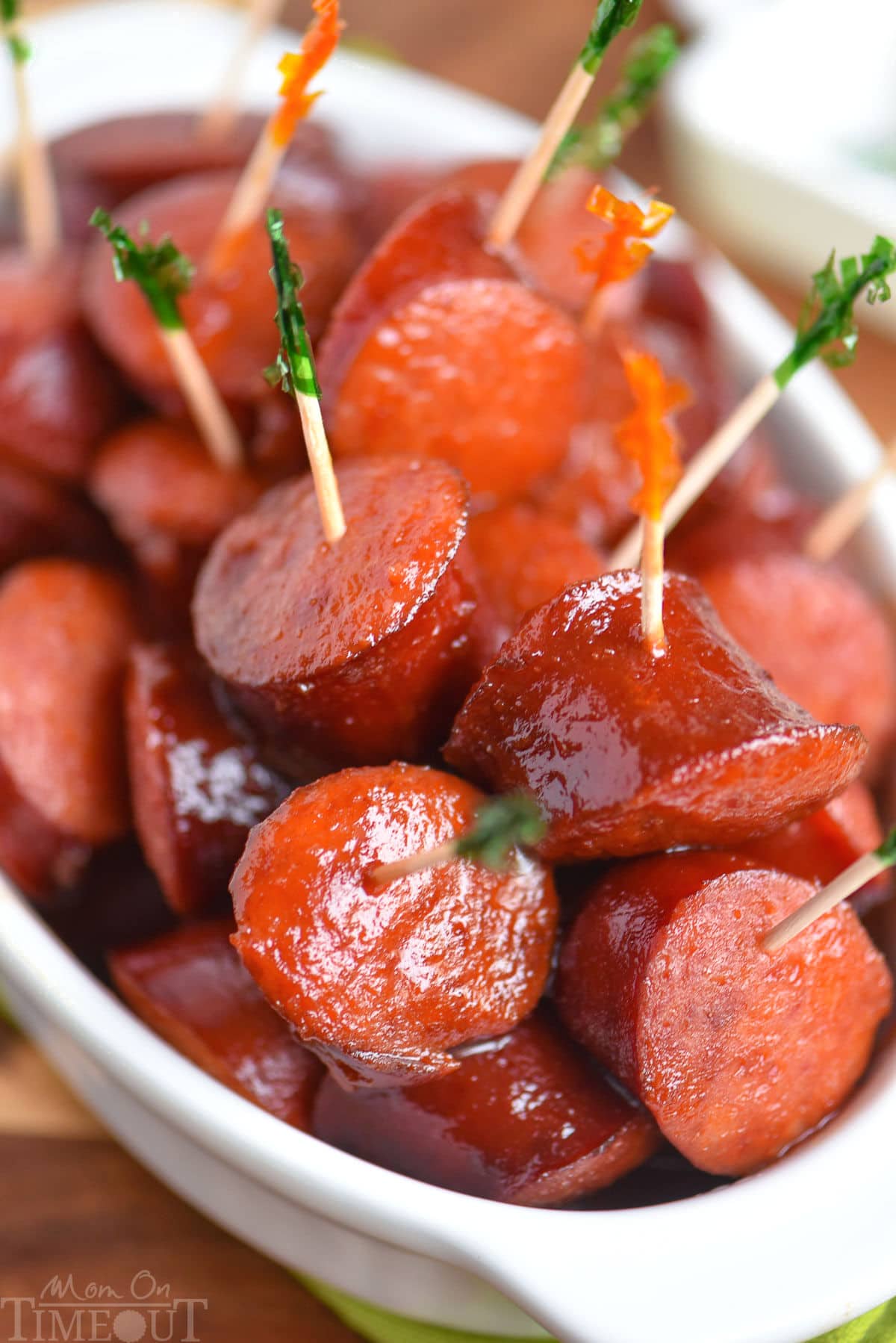 Pieces of kielbasa have been cooked in a slow cooker in a mouthwatering sauce and are shown in a white bowl with toothpicks in them.