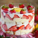 front view of strawberry and lemon trifle layered in a clear glass trifle dish. Topped with fresh strawberry and lemon slice.