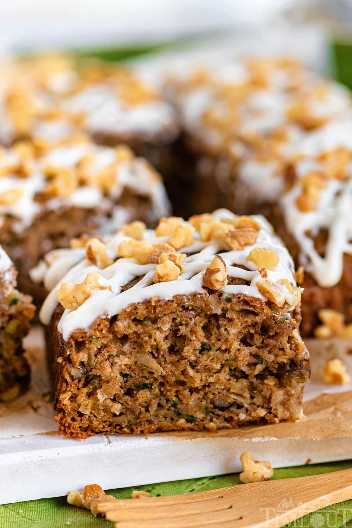 front view of a piece of zucchini snack cake on a board. Cake is frosting with cream cheese icing and topped with walnuts.