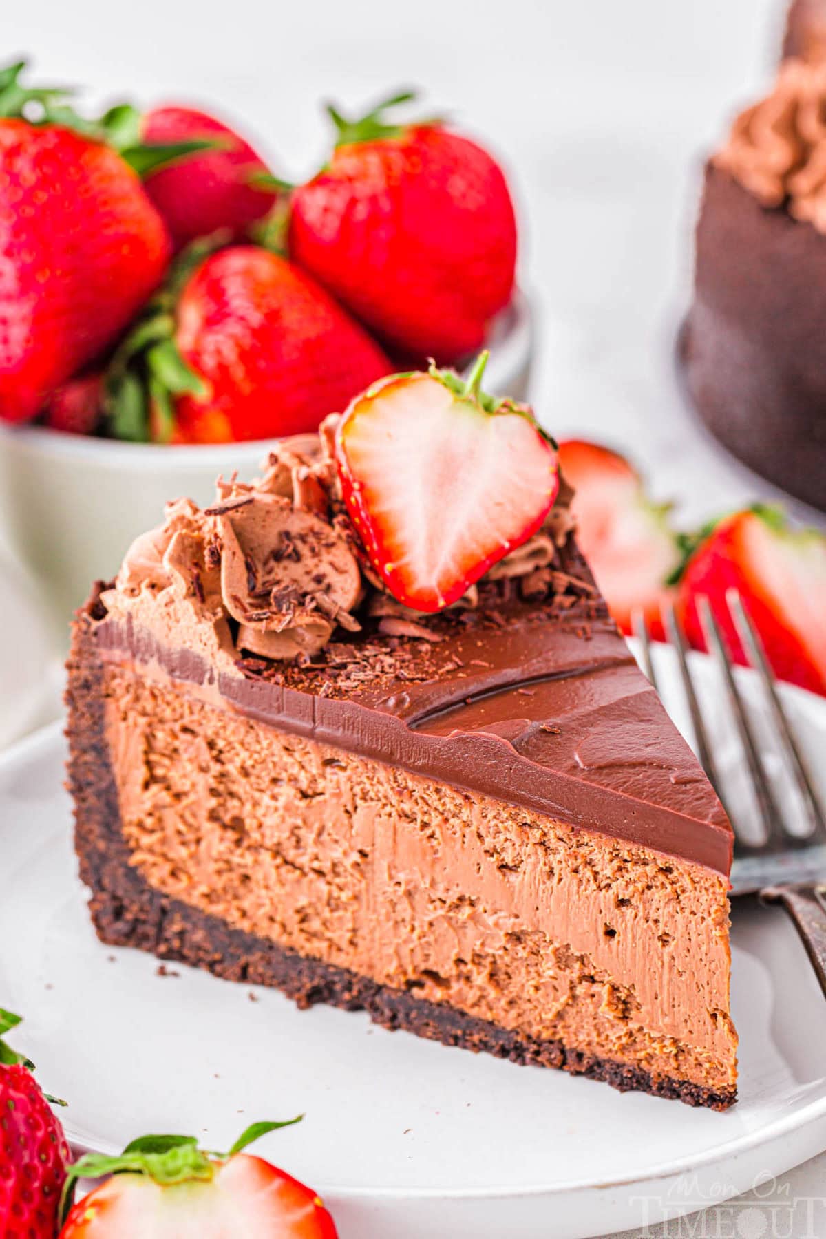 Slice of decadent chocolate cheesecake with an oreo crust and rich chocolate ganache on white round plate. Topped with chocolate shavings and fresh strawberries.