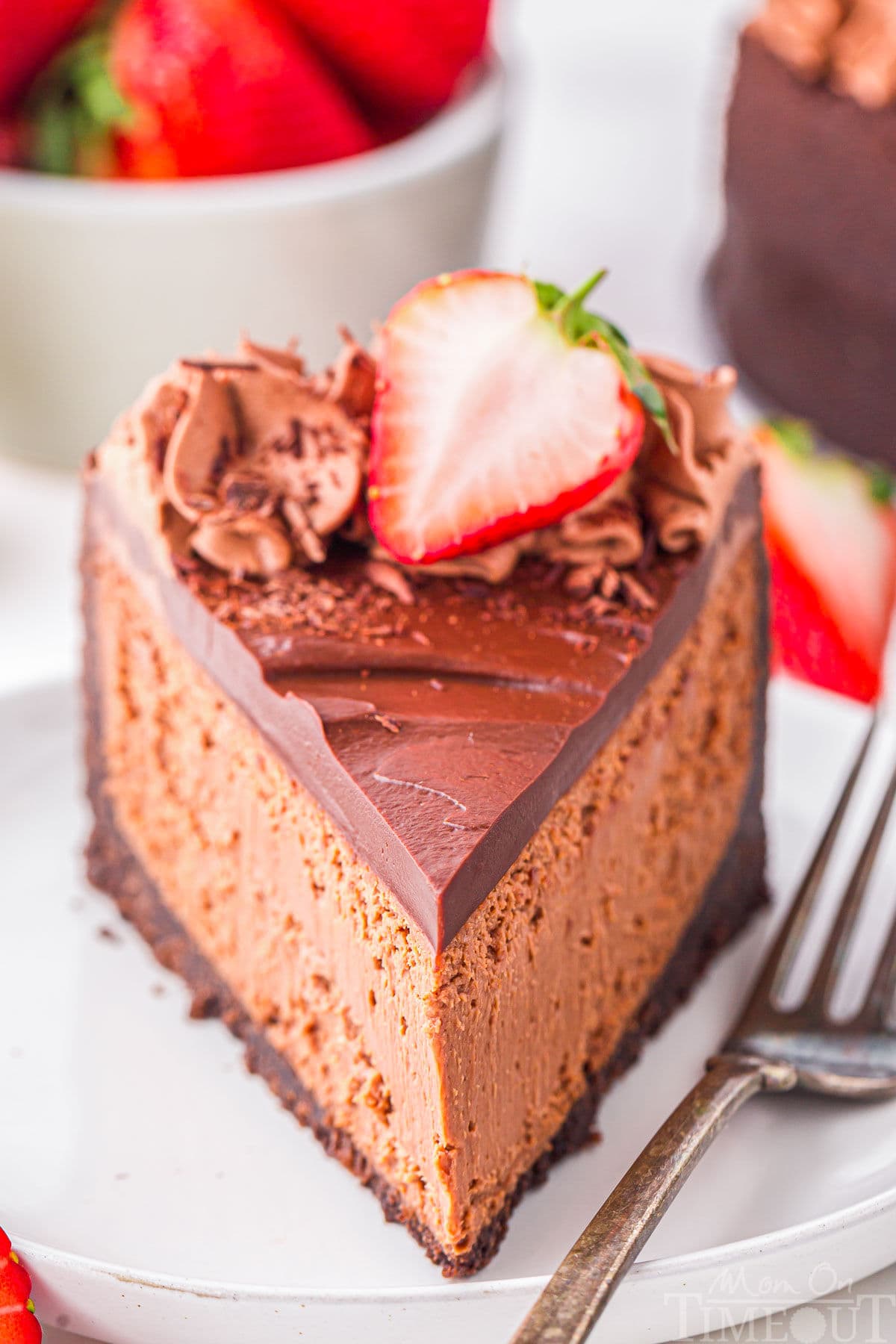 Slice of decadent chocolate cheesecake with an oreo crust and rich chocolate ganache on white round plate. Topped with chocolate shavings and fresh strawberries. Strawberries and more cheesecake can be seen in the background.