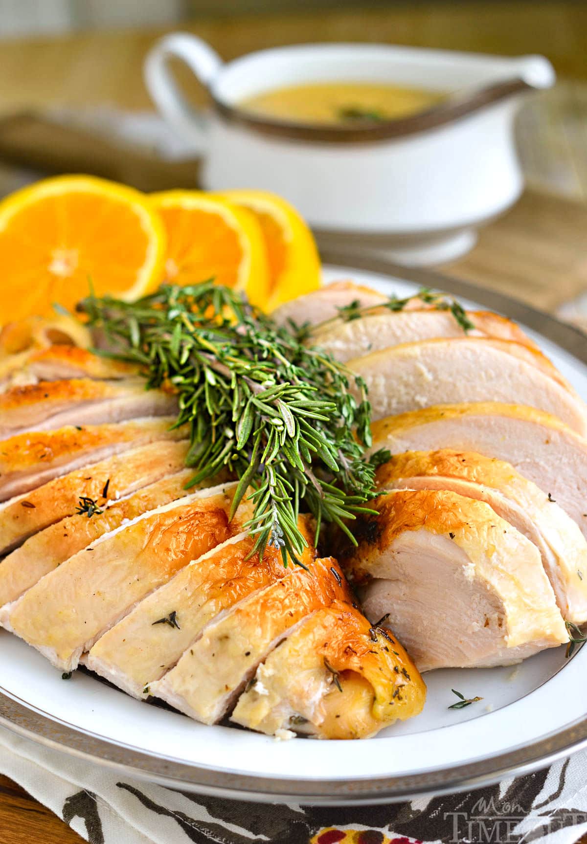 Turkey breast on white platter with rosemary and orange slices already sliced and ready to be served. Turkey gravy can be seen in the background.