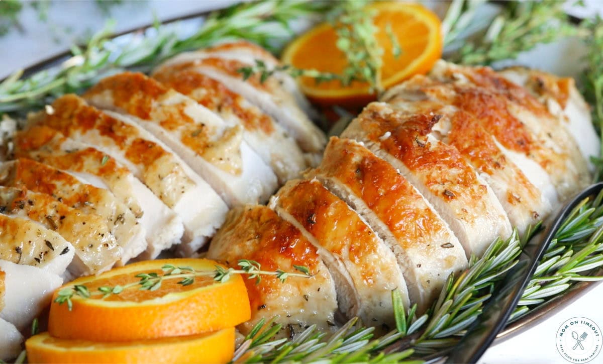 Turkey breast in crockpot cooked to perfect, broiled for crispy skin, sliced and sitting on a serving plate with fresh herbs and orange slices.