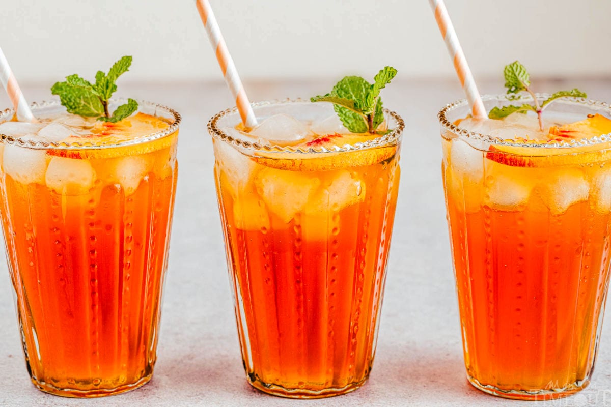 three glasses of peach iced tea in a line garnished with mint sprigs and slices of peach.