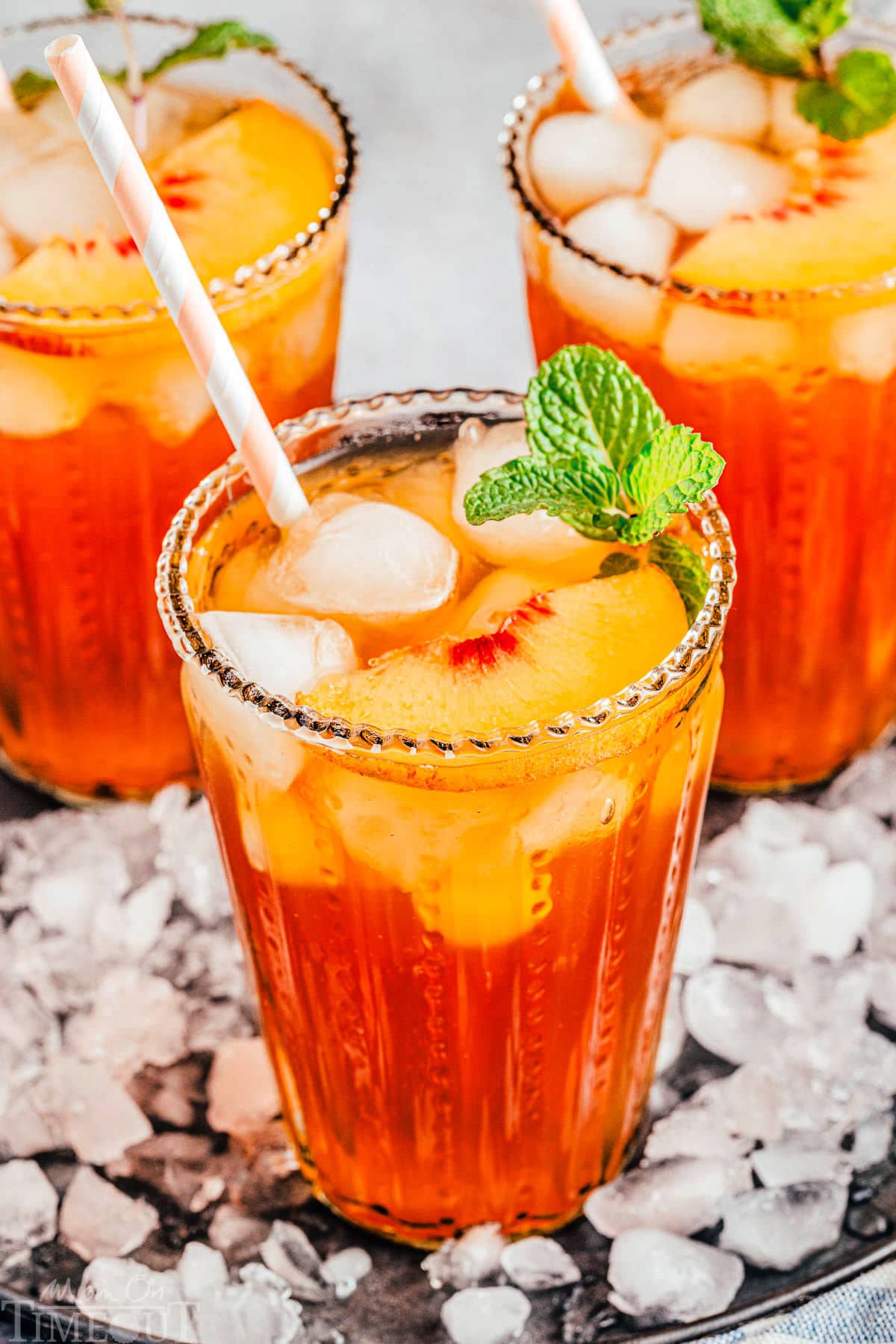 three tall glasses filled with homemade peach iced tea and topped with fresh peach slices and a sprig of mint. glasses are sitting on a tray covered with ice.
