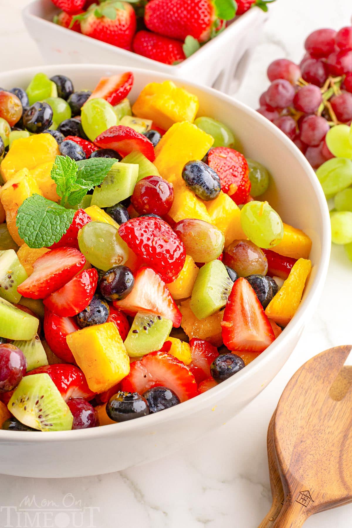 A white bowl of fruit salad with fruit in the background.