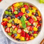 A top down view of fruit salad in a white bowl with wood salad tongs sticking out of it.
