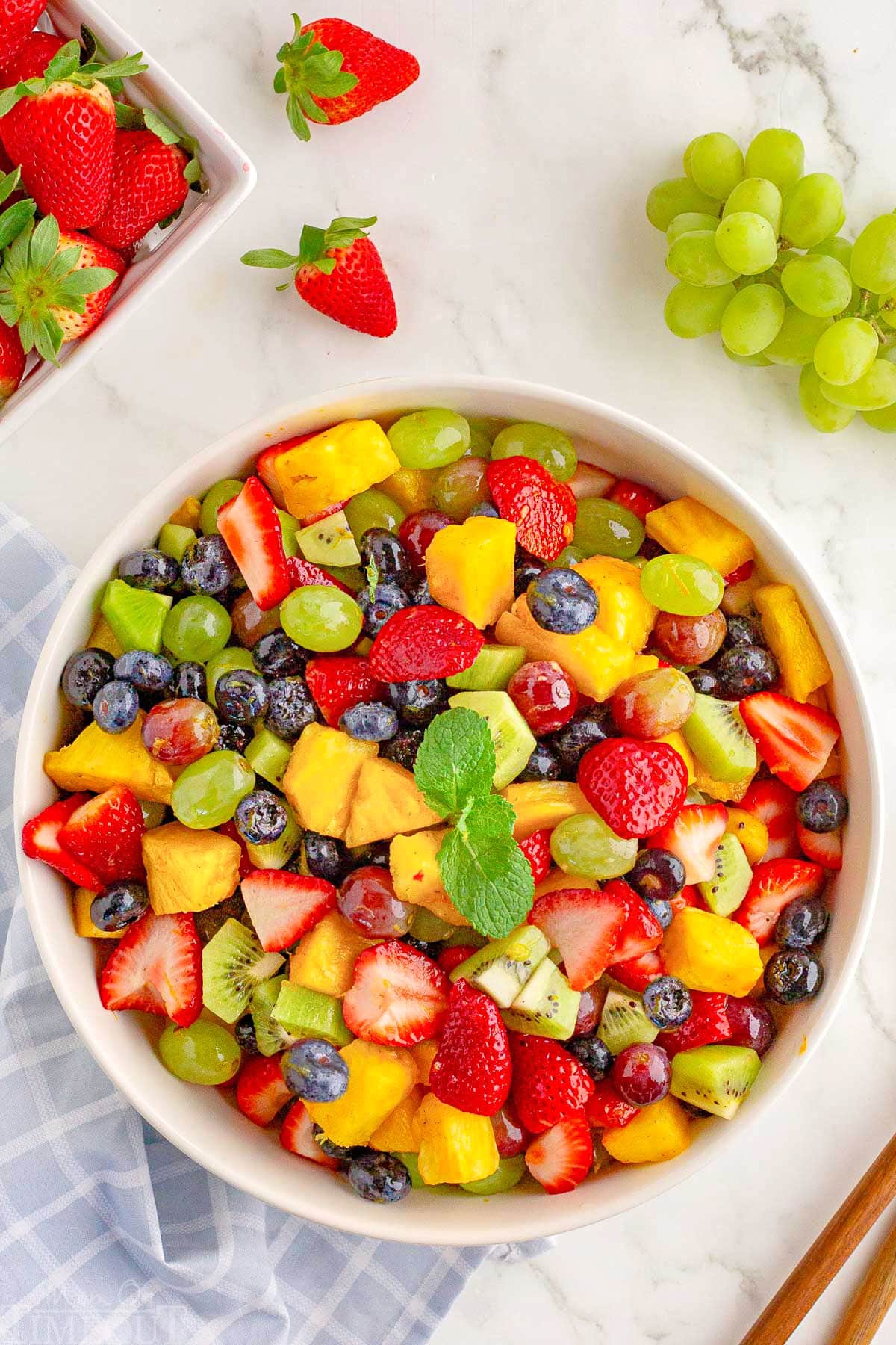 top down shot of fresh fruit salad in large white serving bowl topped with a sprig of mint. Green grapes and strawberries are scattered around the outside of thr bowl.