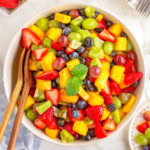 A top down view of fruit salad in a white bowl with wood salad tongs sticking out of it.