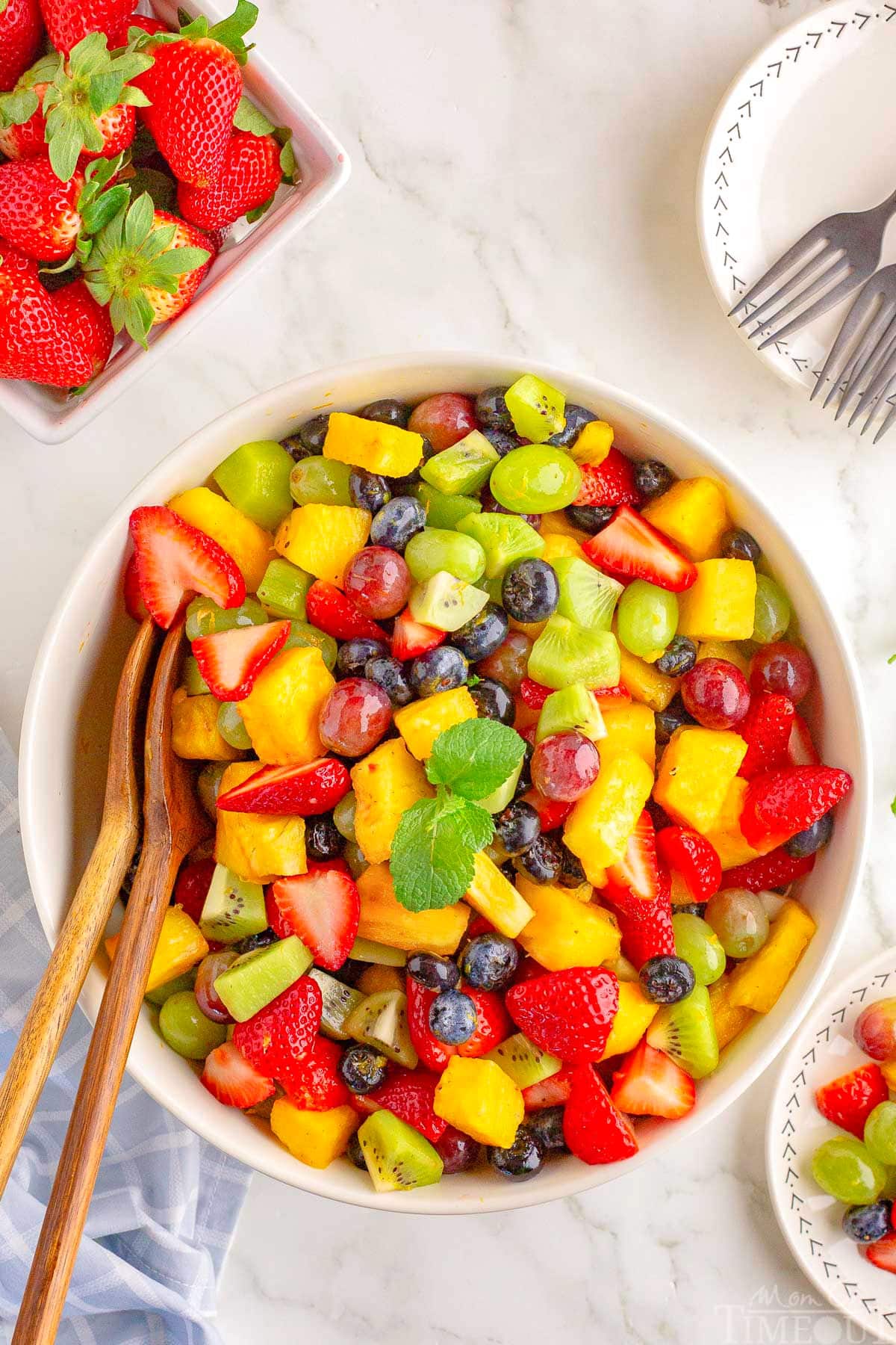 A top down view of a bowl of fruit salad with wood salad tongs sticking out of it.
