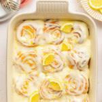 An overhead view of a pan of lemon sweet rolls topped with a lemon cream cheese glaze, lemon slices and lemon zest. Baking dish is white.