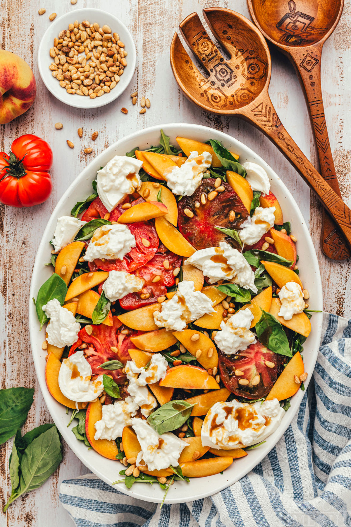 fresh peach salad made with burrata and heirloom tomatoes all topped with a balsamic vinaigrette. Salad is served on a white oval platter and is topped with pine nuts.