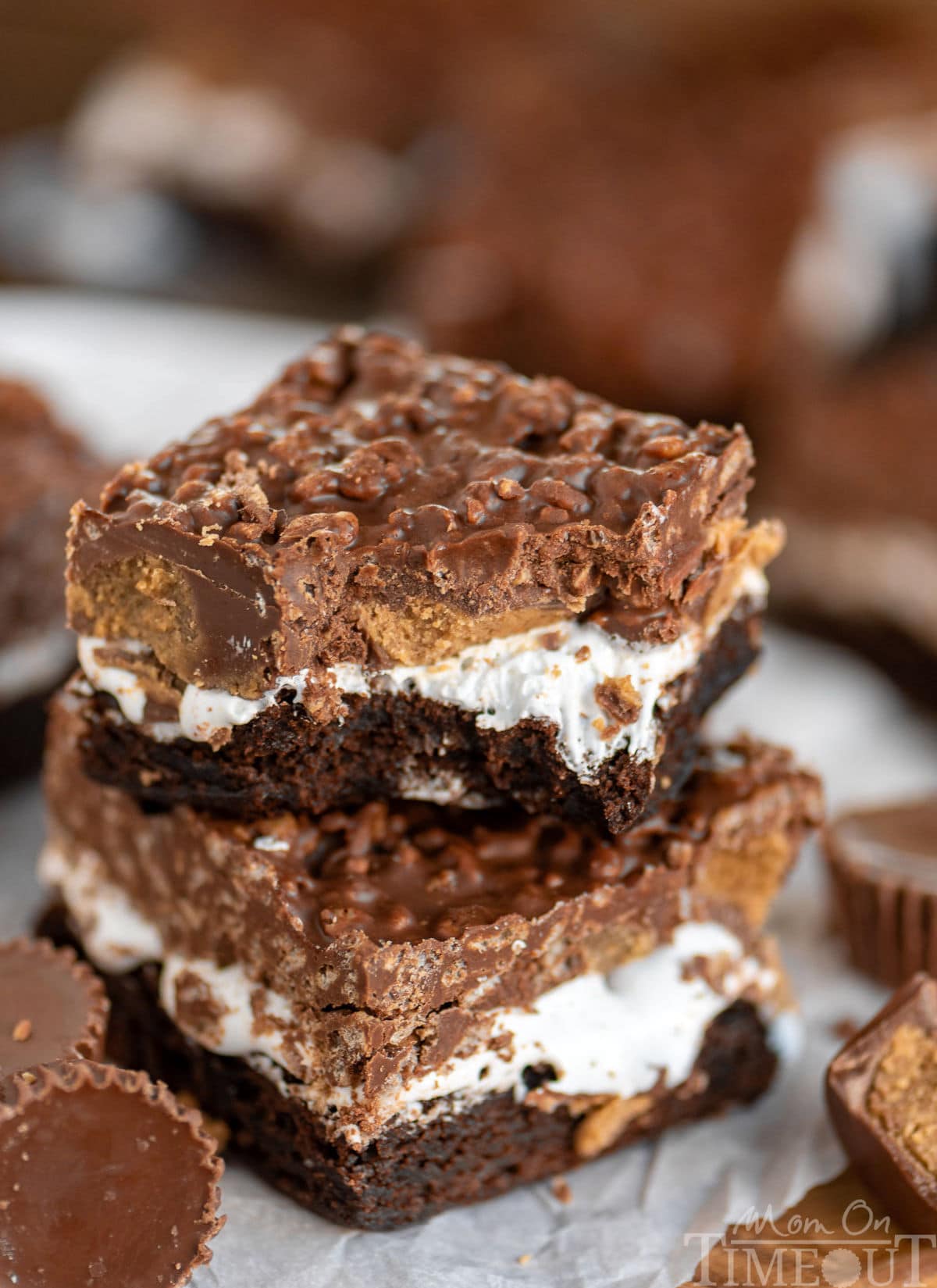 Two brownies with multiple layers including marshmallow, Reese's candy, and a crunch peanut butter chocolate layer stacked on each other. The top brownie has a bite taken out of it.