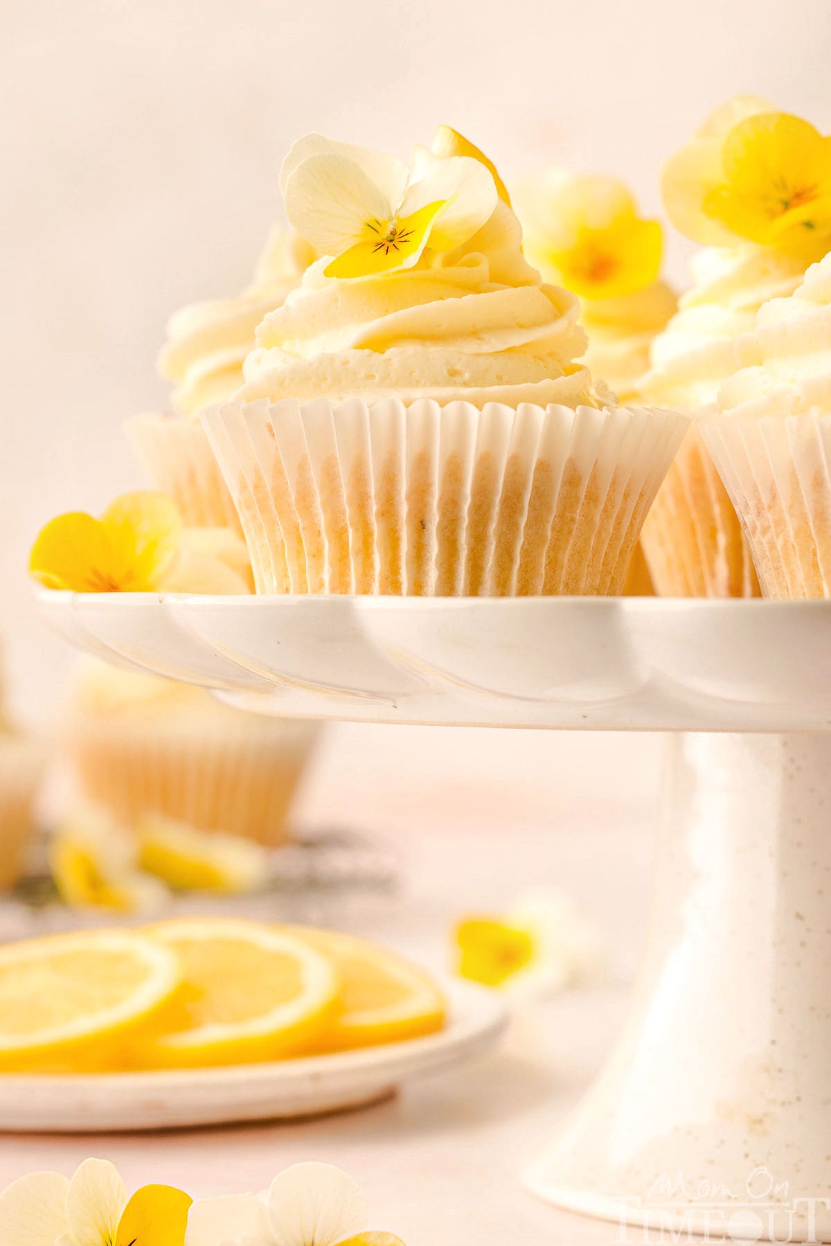 Lemon cupcakes on a scalloped edge white cake stand. Lemon slices and more cupcakes can be seen in the background.