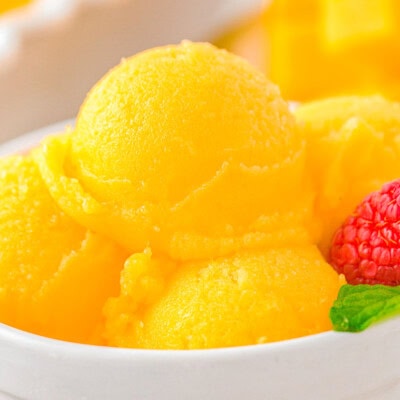 Scoops of mango sorbet in a white bowl topped with fresh mint and raspberries.