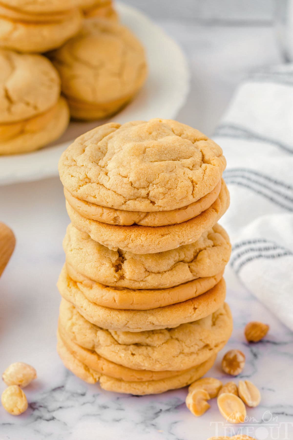 Three peanut butter sandwich cookies stacked on each other on a marble surface. A plate of more cookies can be seen in the background.