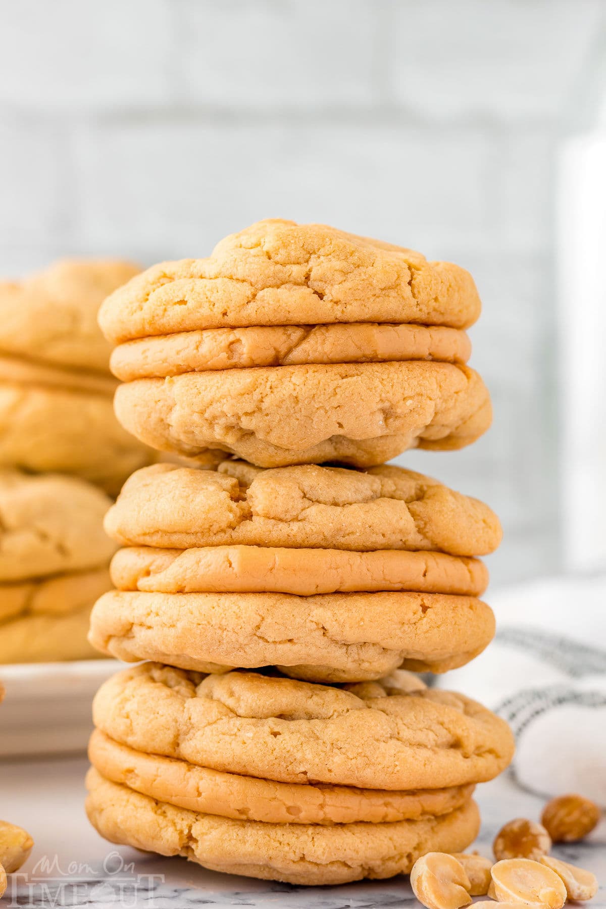 Three peanut butter sandwich cookies stacked on each other on a marble surface. A plate of more cookies can be seen in the background.