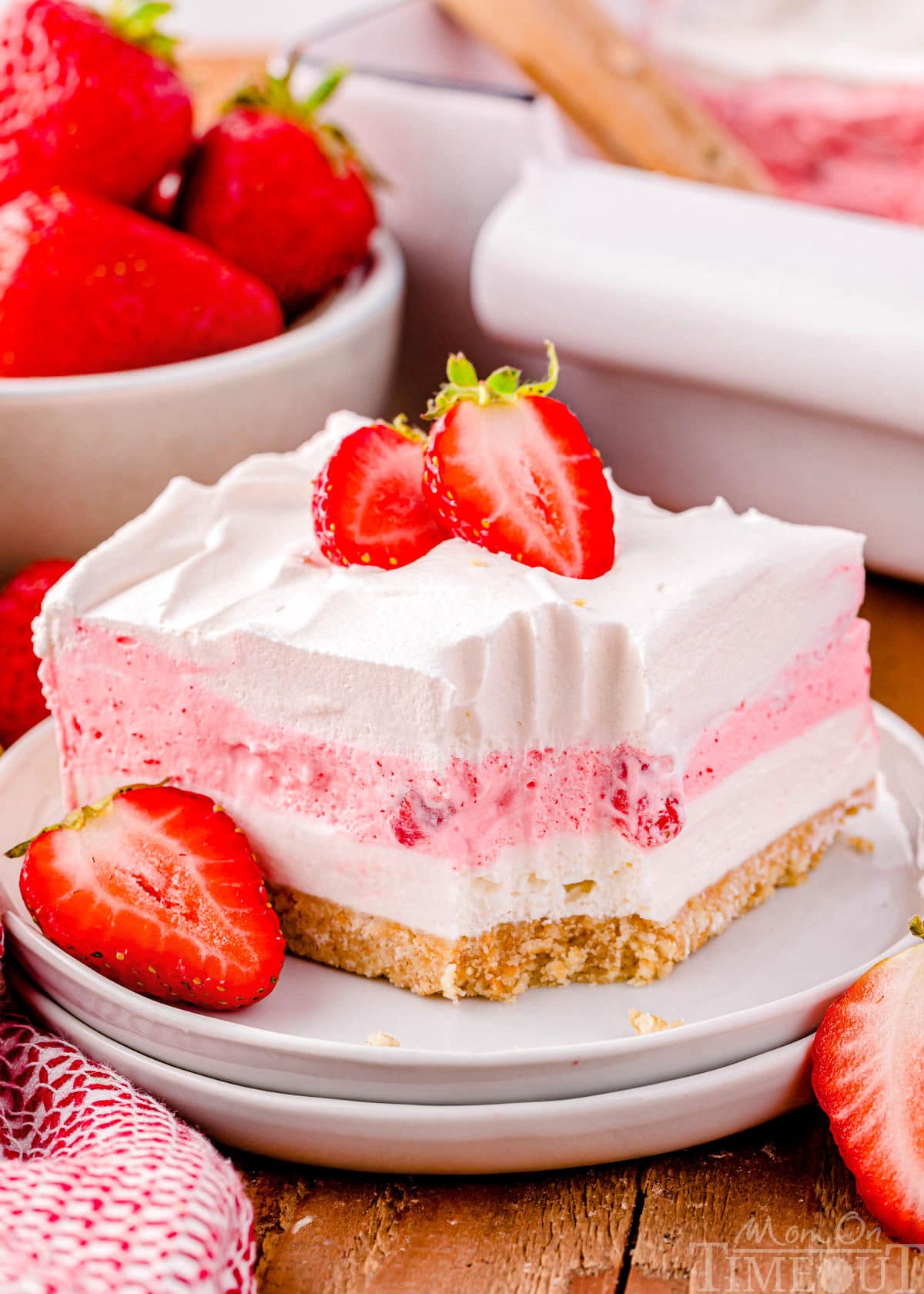 A square of strawberry dessert lasagna on a round white plate topped with whipped cream and fresh strawberries. A bowl of strawberries can be seen in the background.