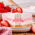 A slice of strawberry lasagna on a white plate topped with fresh strawberries. The baking dish and a bowl of strawberries can be seen in the background.