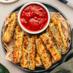 Top down view of air fryer zucchini on a metal plate with a small bowl of marinara to dip the fries in.