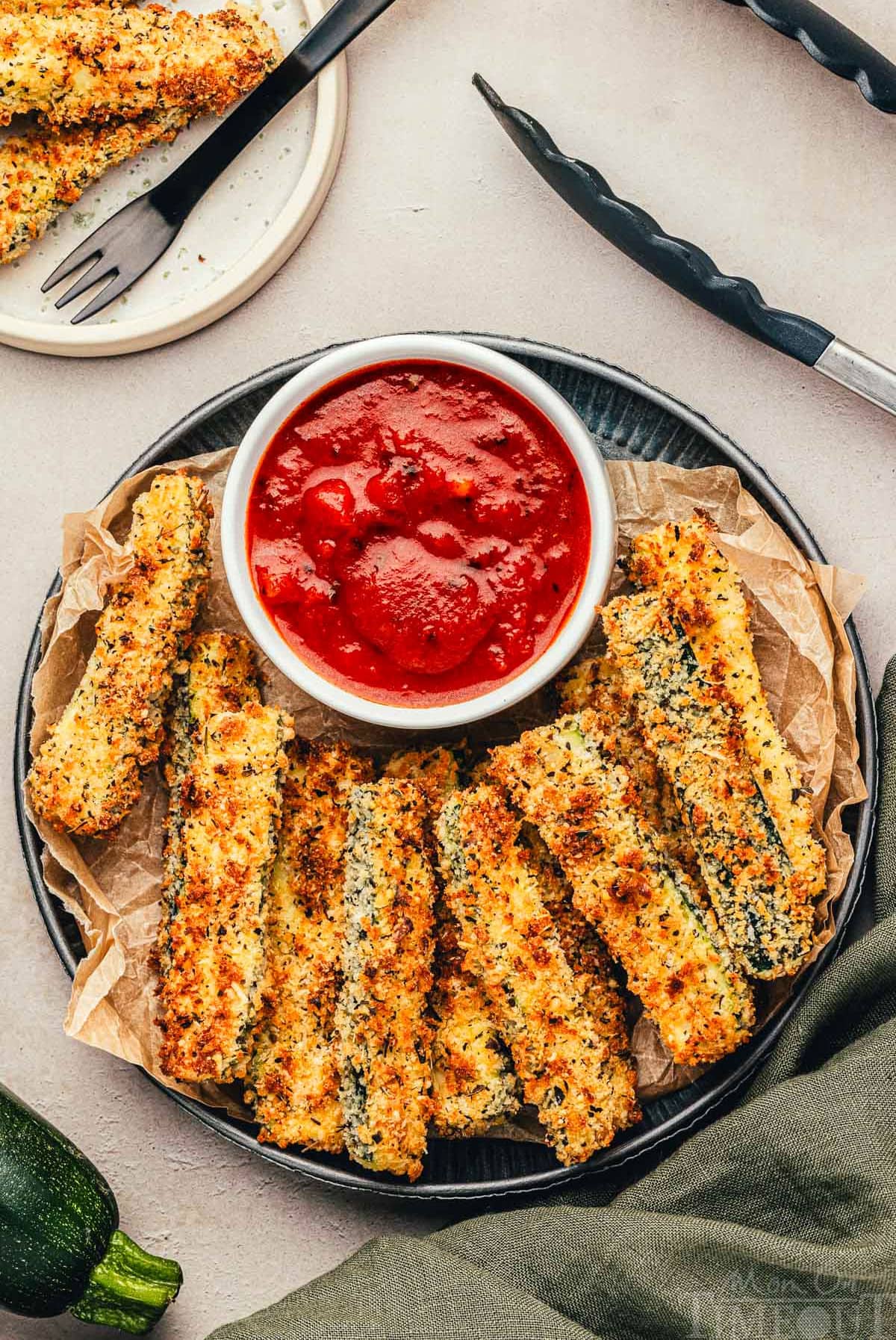 Top down view of air fryer zucchini on a metal plate with a small bowl of marinara to dip the fries in. 