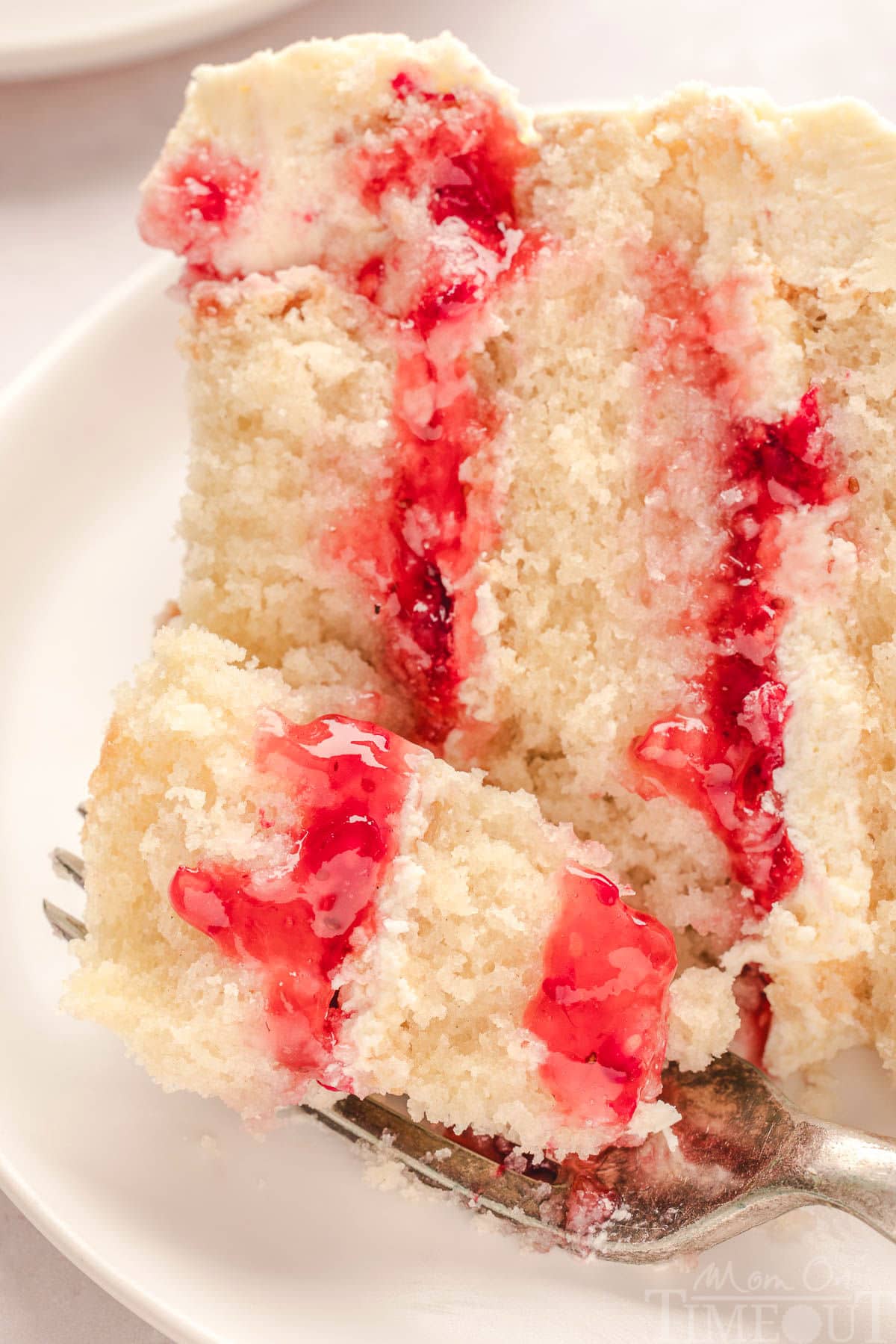 slice of raspberry white chocolate cake on white round plate. Fork has cut off a bite of the cake.