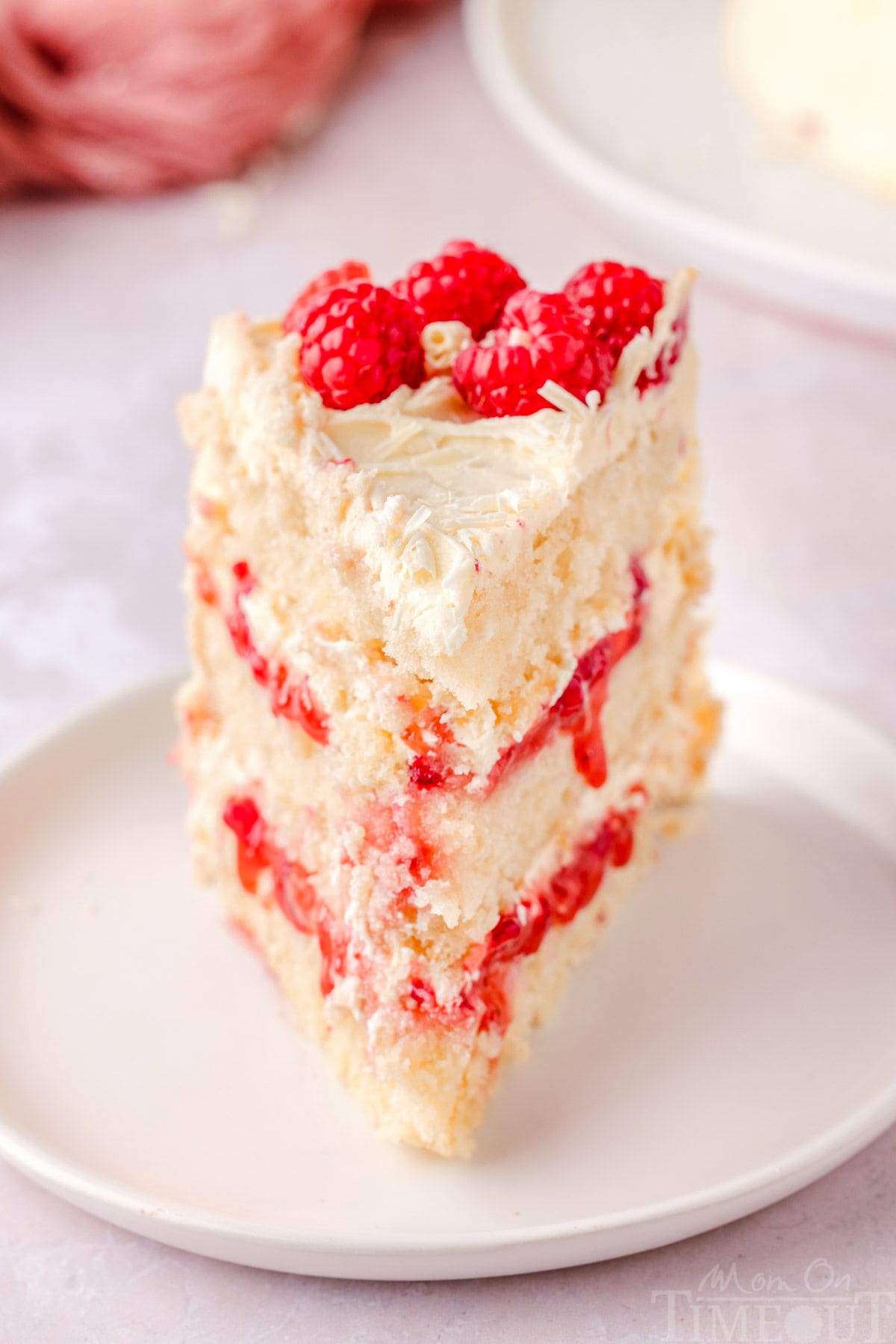 Slice of white chocolate raspberry cake on a white round plate topped with fresh raspberries and white chocolate curls.