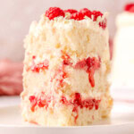 Slice of white chocolate raspberry cake on a white round plate topped with fresh raspberries and white chocolate curls.