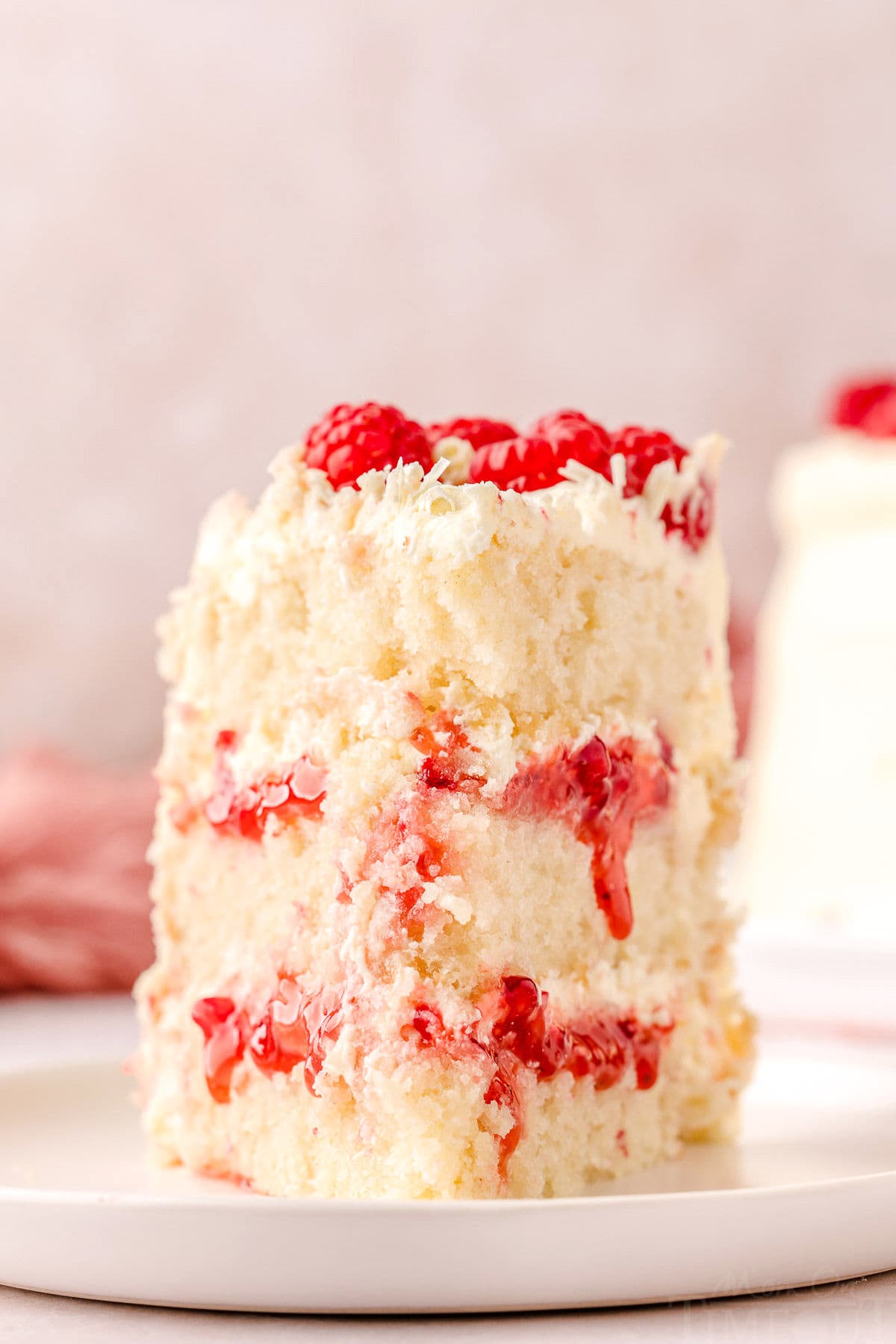 Slice of white chocolate raspberry cake on a white round plate topped with fresh raspberries and white chocolate curls.