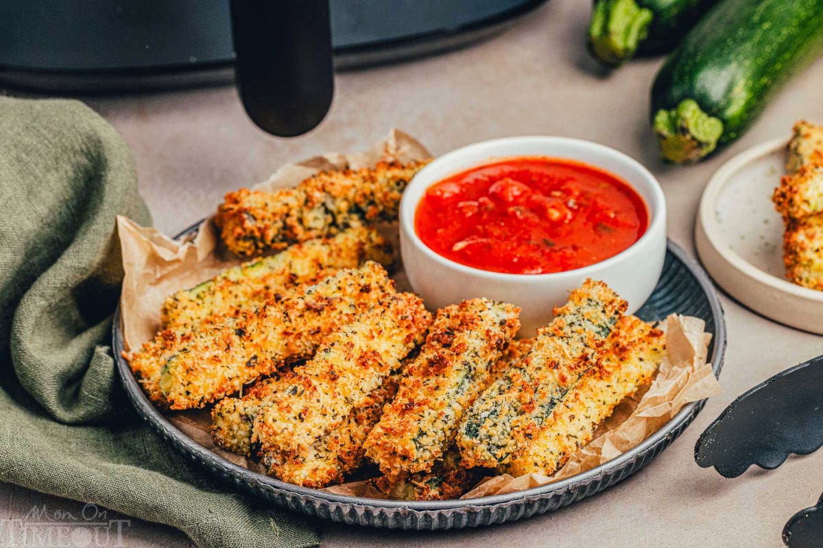 Wide shot of zucchini fries on a metal plate with black air fryer in the background.