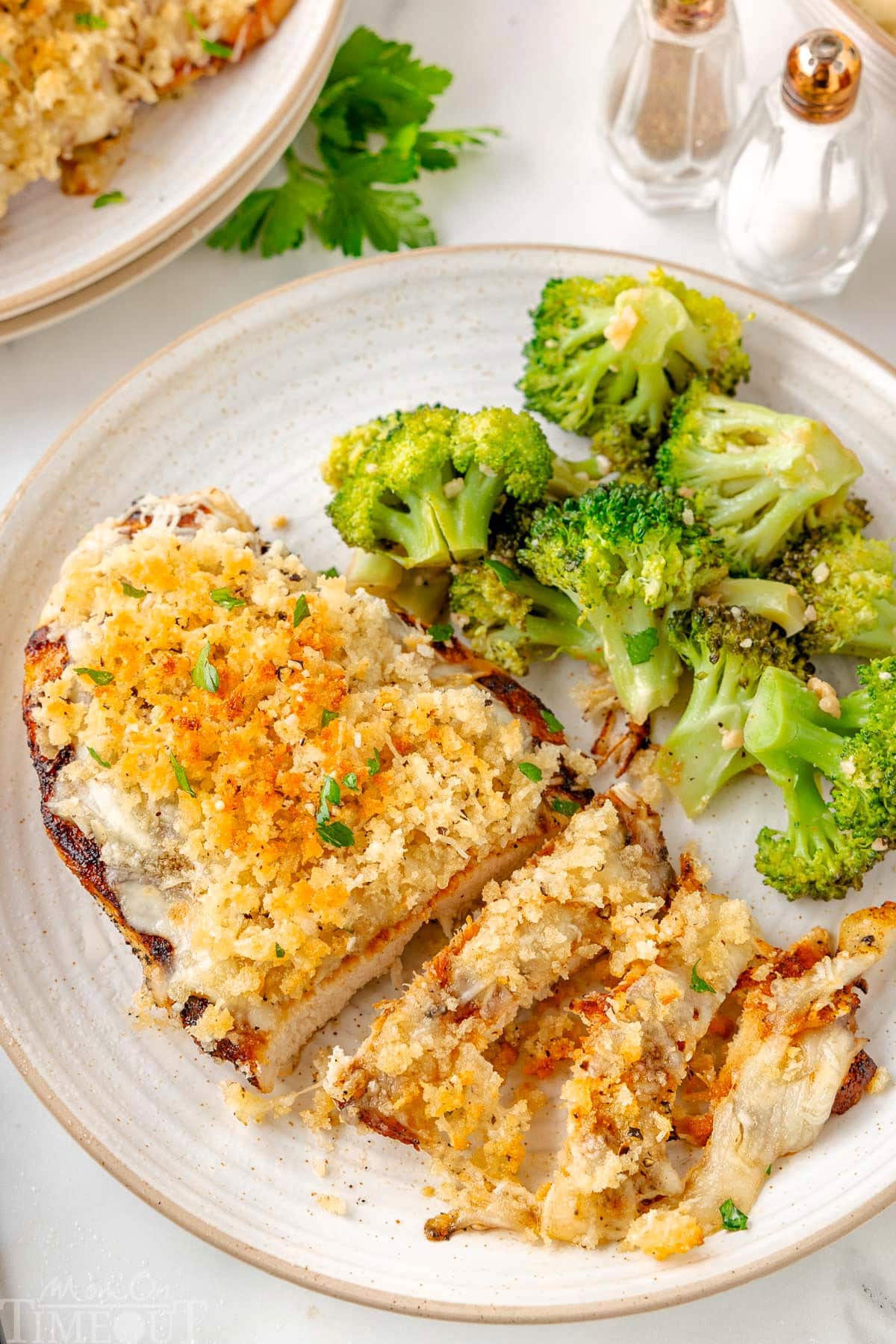 Top down view of Longhorn Parmesan Crusted Chicken plated with roasted broccoli on a round white plate. The crispy Parmesan and Panko topping is clearly shown and a few strips of chicken have been cut off the breast.