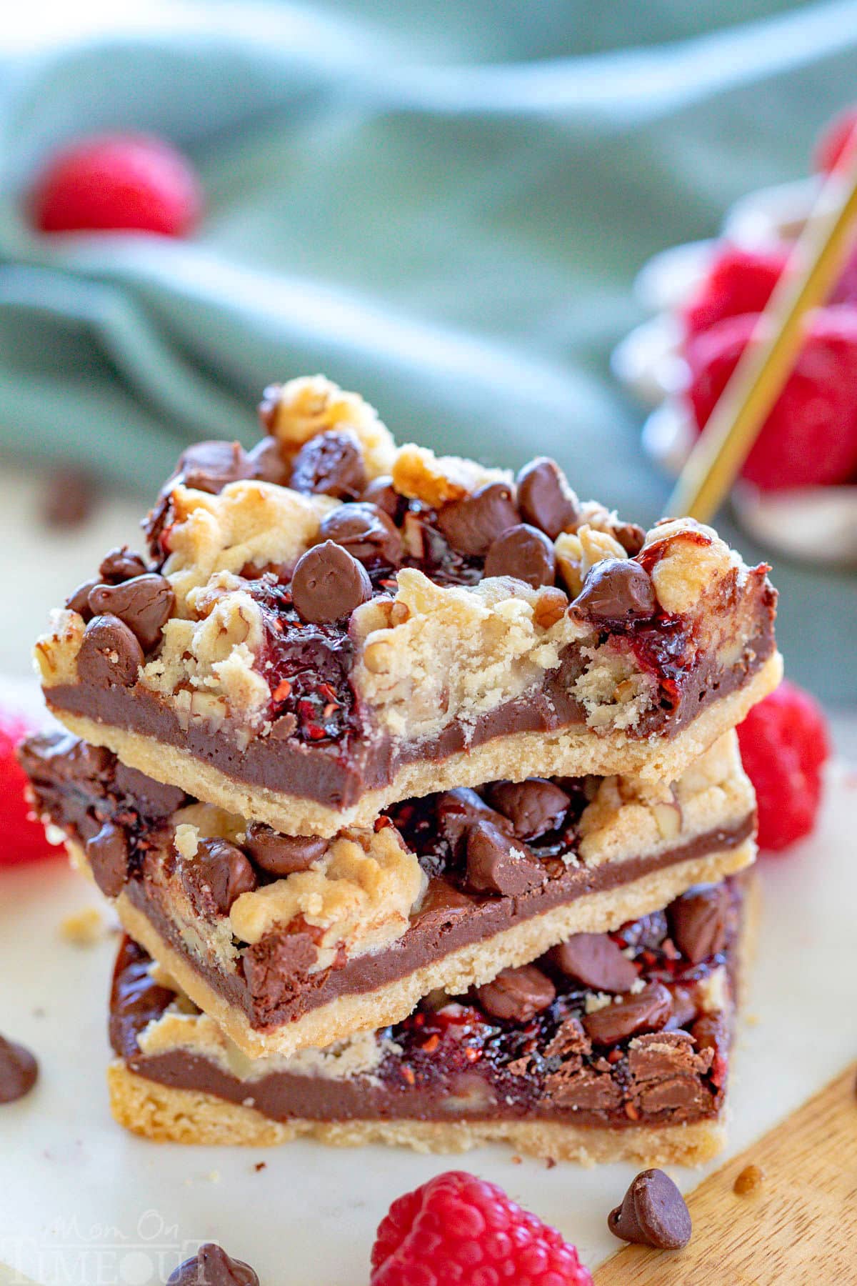 Three chocolate raspberry crumb bars stacked on a white marble board. Fresh raspberries can be seen in a white bowl in the background. The top bar has a bite taken from it.