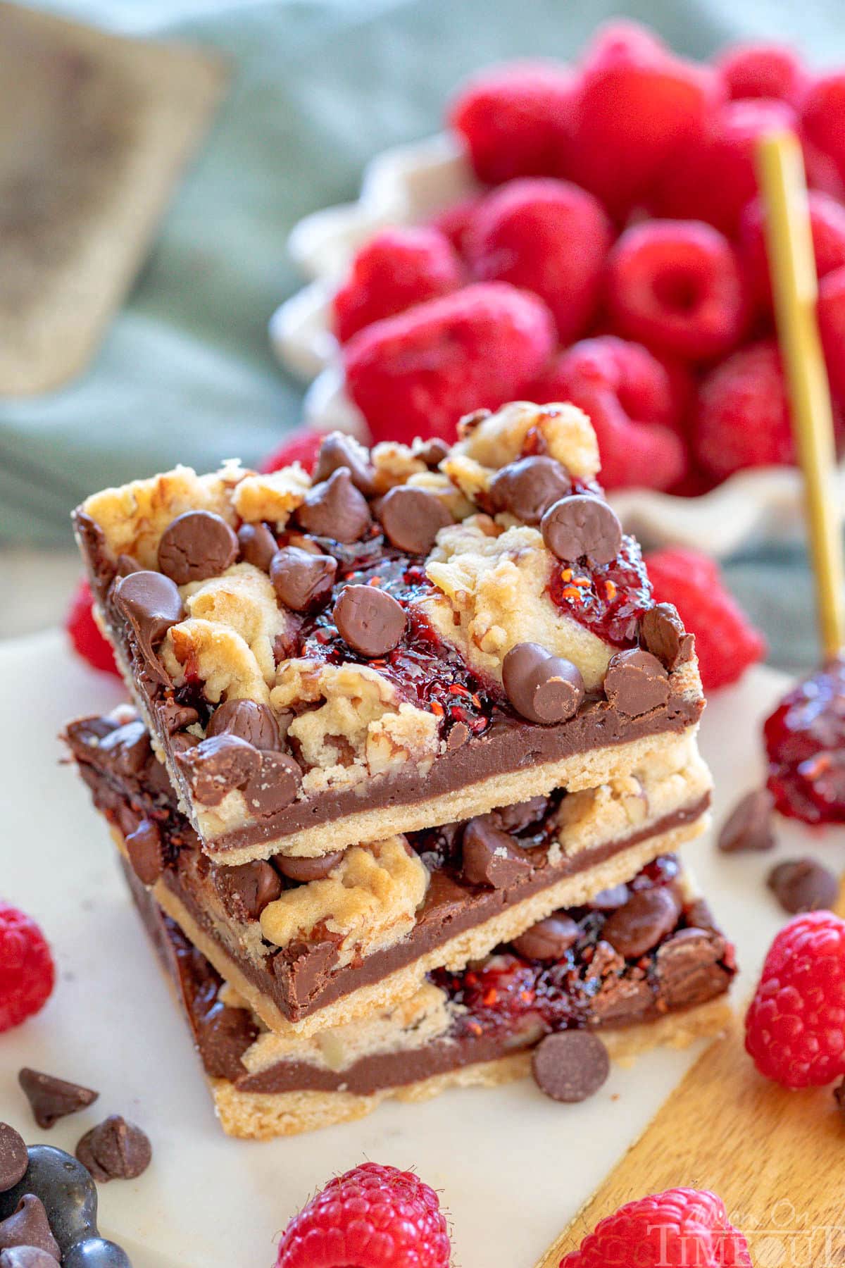 Three chocolate raspberry crumb bars stacked on a white marble board. Fresh raspberries can be seen in a white bowl in the background.