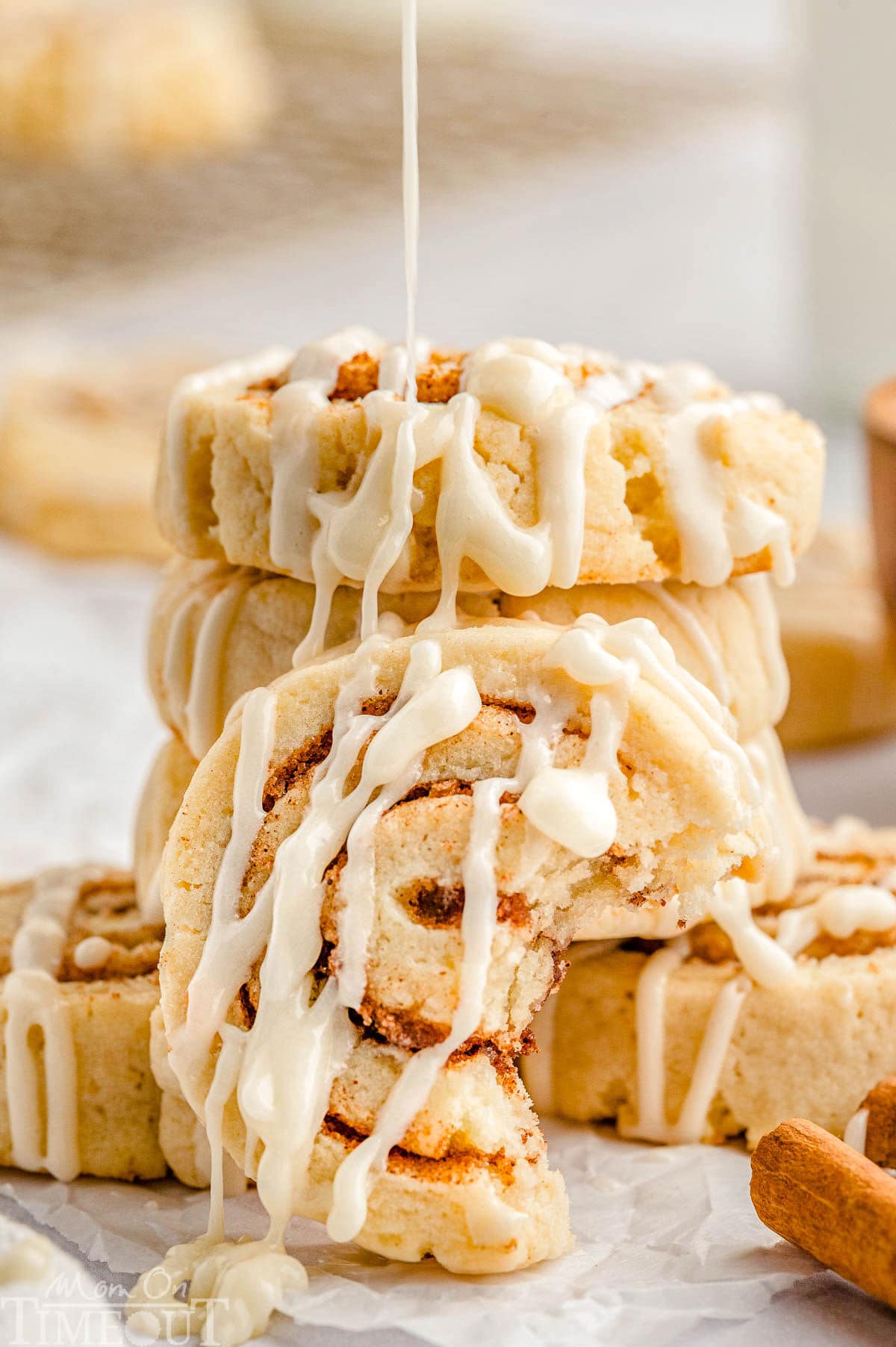 Cinnamon Roll Cookies with icing in a stack. One cookie is leaning on the stack with a bite taken. Icing is being drizzled onto the cookies.