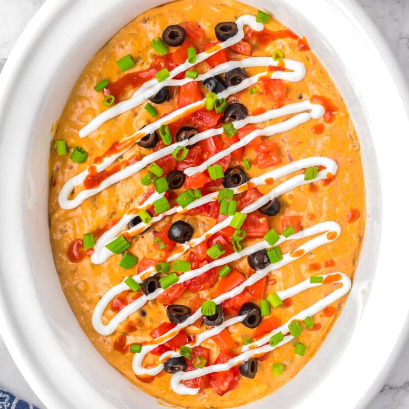 Top down view of crockpot taco dip in a white crockpot. The dip has been topped with lots of taco toppings.